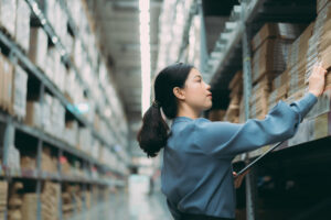 Young smart Asian business working woman thinking while using digital tablet to check goods on shelves for product management in warehouse, Logistics business planning concept with copy space