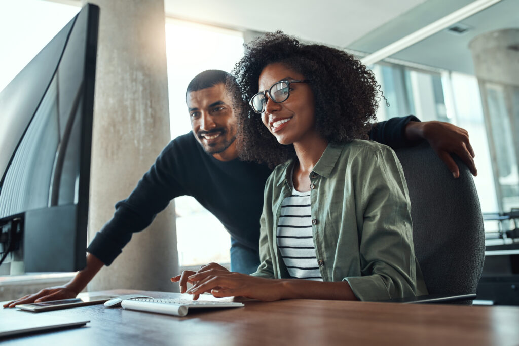 An african young businesswoman sitting at her desk working on computer with male colleague standing by to depict a Data Commerce team